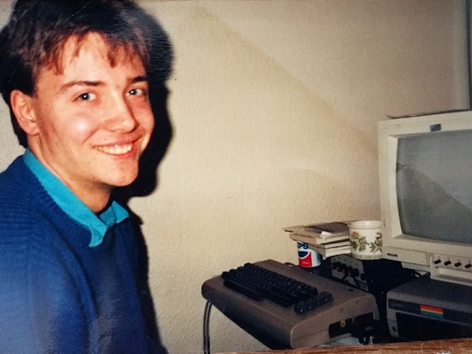 A photograph of a young Simon Pick with his Commodore 64. Pick beams a winning smile, facing the machine.