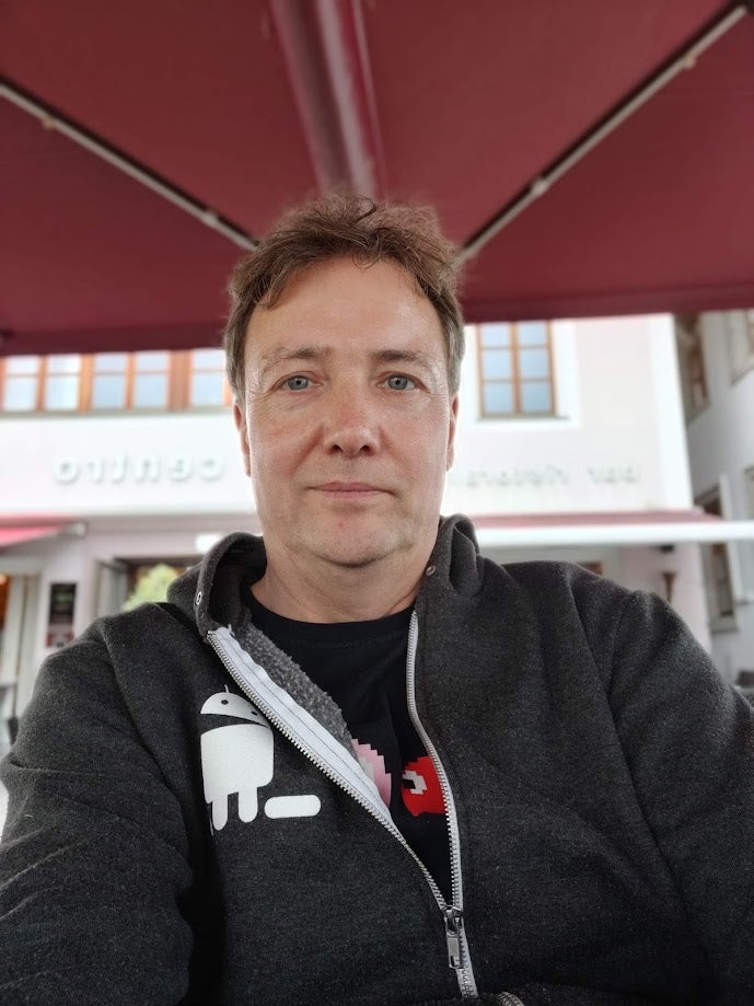 A selfie of Simon Pick taken recently. He's sits under parasol in a city somewhere, wearing a game-related hoodie.