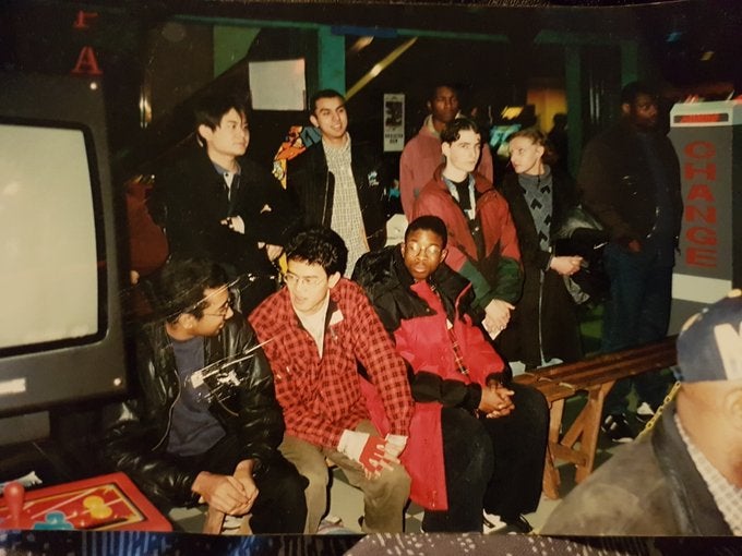 Scanned photo of Ryan Hart and friends at a gaming event in the mid 90s. Hart wears a large red overcoat and sits on a basic wooden bench by a large CRT monitor.