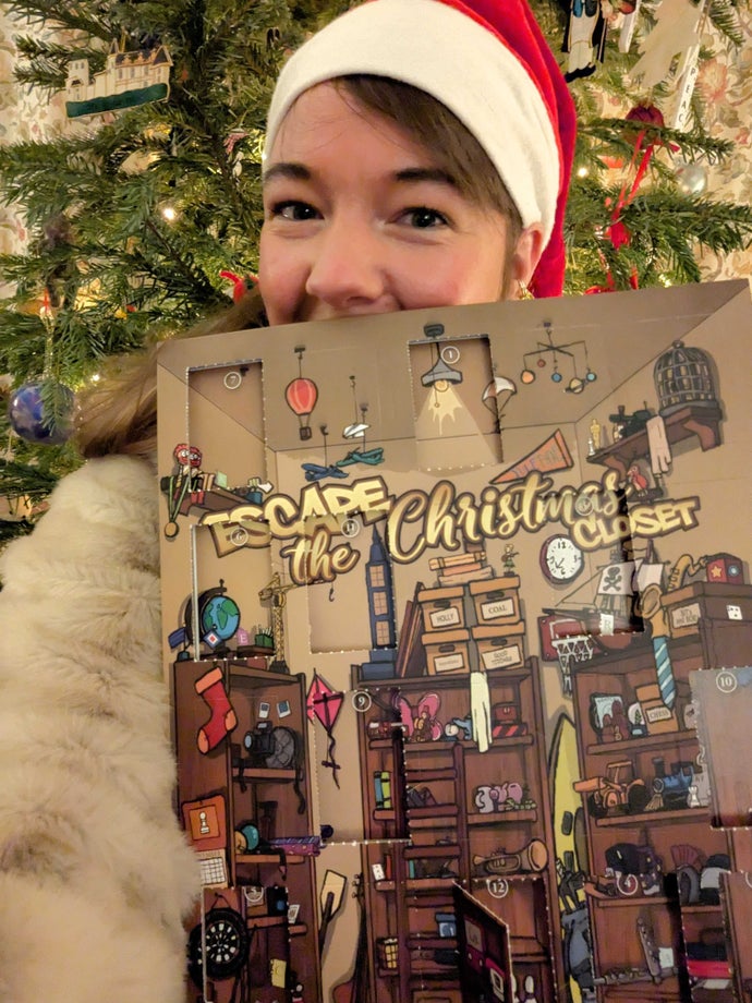 A photo of Eurogamer reporter Victoria Kennedy holding up her escape room advent calendar. She's wearing a Santa hat and there's a Christmas tree behind her.