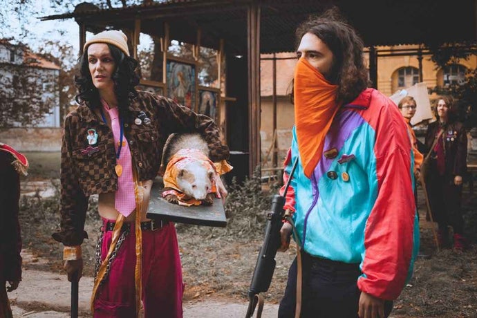 Two costumed performers at a Disco Elysium LARP. One is holding a replica gun with an orange bandana across his face. The other is holding a taxidermy on a wooden board.